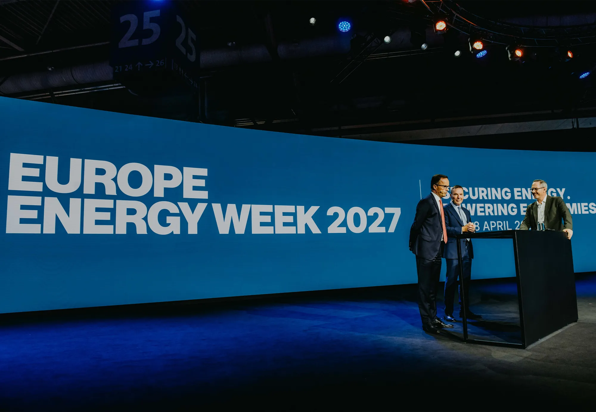 europe energy week logo on large wide screen, used as the backdrop for a stage. 3 men stand at a podium on the stage